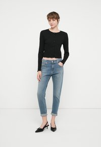 Črn skrajšan kardigan z dolgimi rokavi in gumbi, v kombinaciji s svetlo modrimi ozkimi kavbojkami in črnimi čevlji na peto.