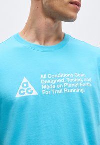 Άνδρας φοράει ανοιχτό μπλε μπλουζάκι με λευκό κείμενο «All Conditions Gear. Σχεδιασμένο, δοκιμασμένο και κατασκευασμένο στον πλανήτη Γη. Για trail running.» και λογότυπο ACG.