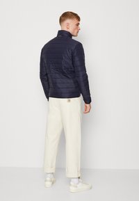 Giacca puffer blu navy con cuciture orizzontali, colletto alto e dettaglio del logo sul retro, abbinata a pantaloni color crema e sneaker bianche.
