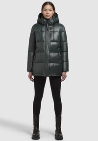 Vrouw in een donkergroene gewatteerde jas met capuchon, zwarte leggings en zwarte enkellaarsjes, staande tegen een effen lichte achtergrond.