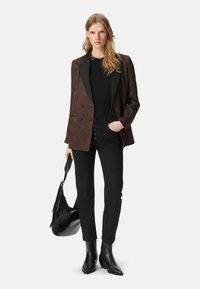 Blazer marrone con motivo e rever neri, camicia nera, jeans neri a vita alta con dettagli in bottone, e stivali neri alla caviglia, mentre tiene una borsa nera.