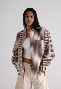 Stribet skjorte i beige og burgunder med en enkelt brystlomme og broderidezign, parret med lyse bukser.