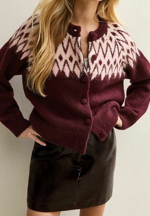 Femme portant un cardigan en maille bordeaux avec des motifs géométriques roses et une mini-jupe en cuir noire, les mains posées sur les hanches.