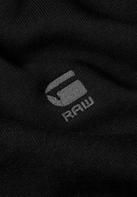 Tejido de punto negro con un logotipo bordado en gris que presenta una 'G' estilizada y la palabra "RAW" debajo, mostrando una textura suave y una costura detallada.