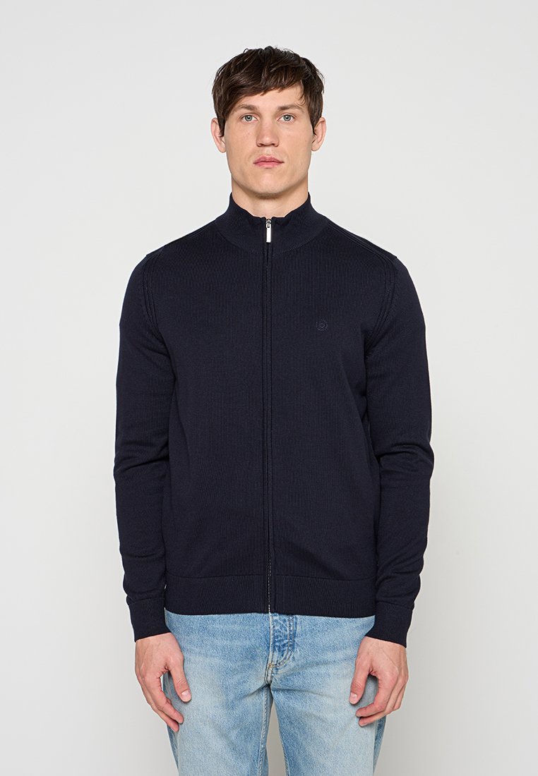 Mann trägt marineblauen Reißverschluss-Pullover und hellblaue Jeans, steht vor einem einfarbigen hellgrauen Hintergrund.