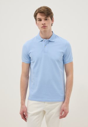 Hombre joven de pie mirando hacia adelante, vistiendo una camisa polo de manga corta azul claro y pantalones blancos sobre un fondo liso.