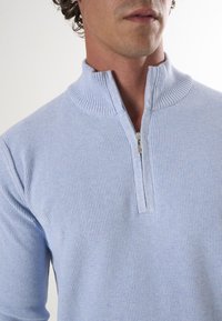 Pull à zip bleu clair avec une texture côtelée, un col montant et un petit détail de zip au niveau du cou.