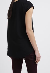 Musta lyhythihainen paita, jossa on pyöristetty helma ja sileä ihotuntuma, yhdistettynä maroonivärisiin slim-fit-housuihin. Yksinkertainen muotoilu, ilman näkyviä koristeita.