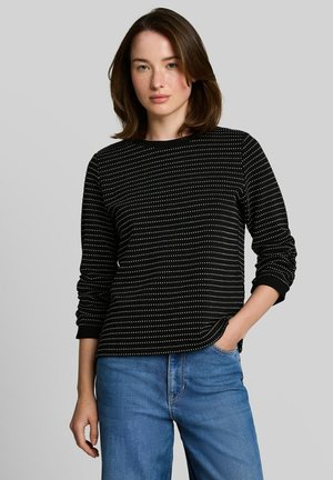 Femme aux cheveux bruns mi-longs porte un haut noir à manches longues avec des pois blancs et un jean bleu, une main dans la poche, fond neutre.