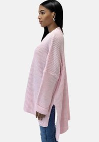 Hellrosa, oversized gestrickter Pullover mit weiten Ärmeln, seitlichen Schlitzen und gerippten Bündchen. Die strukturierte Stoffqualität verleiht dem Design Tiefe.