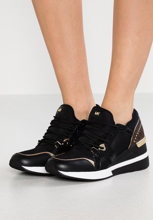 Nogi w czarno-złotych sneakersach Michael Kors z białymi podeszwami, stojące na jasnoszarej podłodze.