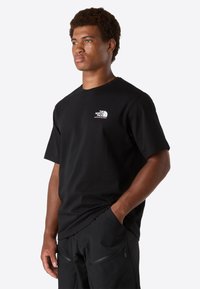 Ung man i svart The North Face t-shirt och svarta byxor, stående med ena handen i fickan och ser lätt åt sidan.