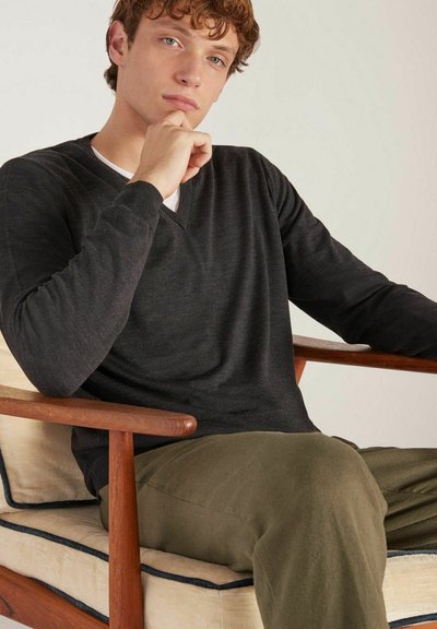 Pull noir en V en tissu doux, cintré à la taille, porté sur une chemise blanche. Mannequin assis sur une chaise en bois avec des coussins de couleur claire.