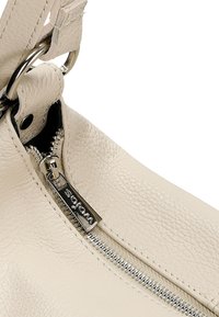 Beige leren handtas met een gestructureerde afwerking, met een zilverachtige rits en een merketiket, in een hoek gepositioneerd om details van de schouderband en ritsafsluiting te tonen.