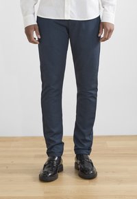 Tmavě modré slim-fit kalhoty vyrobené z denimové látky, s hladkou texturou a minimálním prošíváním, doplněné černými mokasíny.