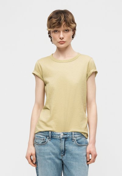 Νεαρός με κοντά κυματιστά μαλλιά φορά ανοιχτό κίτρινο t-shirt με ρολά μανίκια και μπλε τζιν, σε απλό φόντο.