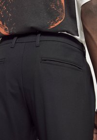 Pantalons noirs sur mesure avec poches passepoilées à l'arrière, portés avec un t-shirt graphique noir et orange rentré à l'intérieur.