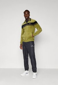 Mees seisab ühe käega taskus, kandes rohelist tõmblukuga jope mustade triipudega, musti spordipükse ja valgeid vabaajajalatsid, taust on ühevärviline.