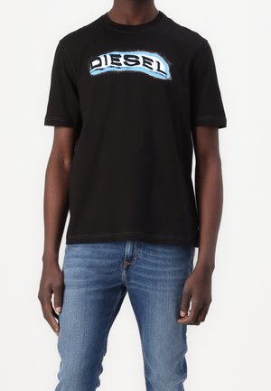 Mann trägt ein schwarzes T-Shirt mit kurzen Ärmeln und dem Schriftzug "DIESEL" in weißer und blauer Schrift, kombiniert mit blauen Jeans, steht vor weißem Hintergrund.