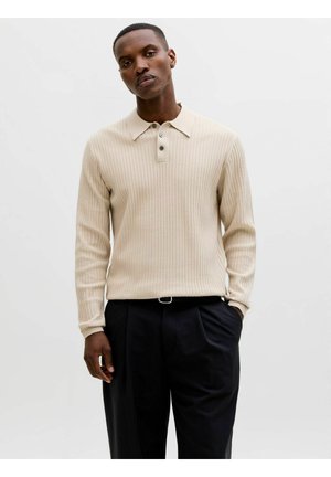 Muž v béžovom rebrovanom polotriku s dlhým rukávom a čiernych nohaviciach stojí pred jednofarebným bielym pozadím.