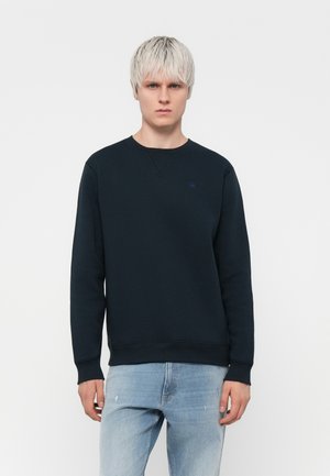 Sweatshirt bleu marine sombre en tissu doux, avec un col rond, des poignets côtelés et un petit logo sur la poitrine. Conçu pour un ajustement décontracté.