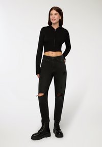 Felpa corta nera a coste con zip frontale, abbinata a jeans neri a vita alta con dettagli strappati sulle ginocchia e stivali neri con lacci spessi.