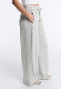 Person trägt hellgraue, weit geschnittene Jogginghose mit Kordelzugbund und seitlichen Taschen, kombiniert mit beigen Sneakern und weißem, kurzen Oberteil.