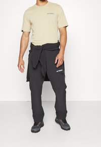 Beige bomuld T-shirt med blåt "Terrex"-logo, kombineret med sorte nylonbukser bundet i taljen og grå vandresko med røde detaljer.