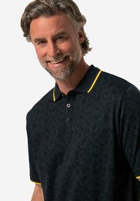 Schwarzes Polohemd mit Paisleymuster, das einen gelben Kragen und Besatz hat, mit drei Knöpfen am Kragen und einem strukturierten Stoff.
