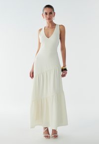 Witte maxi-jurk met lagen en een V-hals, gemaakt van een gestructureerde stof. Voorzien van brede bandjes en een vloeiende onderkant. Draagt bijpassende sandalen met bandjes.