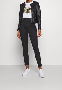 Czarna skórzana kurtka, biały t-shirt z grafiką oraz czarne dopasowane spodnie w połączeniu z białymi tenisówkami. Strój ma swobodny, nowoczesny styl.