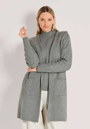 Frau trägt einen grauen gerippten Rollkragenpullover mit einem passenden langen Cardigan, der über strukturierte Taschen verfügt, und weißen Hosen.