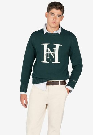 Suéter verde oscuro con un logo y texto blancos, usado sobre una camisa de color azul claro con cuello. Pantalones beige, cinturón marrón y reloj visibles.