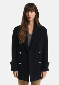 Vrouw met lang bruin haar, gekleed in een zwarte dubbelzijdige jas met gouden knopen en een grijze V-hals trui eronder.