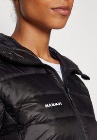 Zwarte pufferjack met capuchon en rits, met een strakke uitstraling, gequiltde textuur en wit "MAMMUT" logo op de borst.
