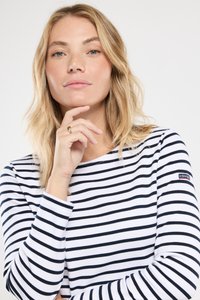 Kvinde med blonde hår iført en hvid og sort stribet langærmet skjorte, hvilende med hagen på hånden med en guldring, neutral udtryk.