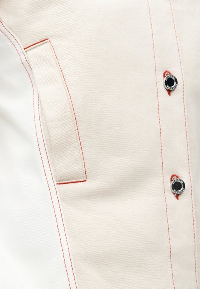 Μπεζ βαμβακερό πουκάμισο με κόκκινες ραφές, με τσέπη πατς και μαύρα μεταλλικά κουμπιά ως διακοσμητικά στοιχεία. Ομαλή υφή, δομημένος σχεδιασμός.