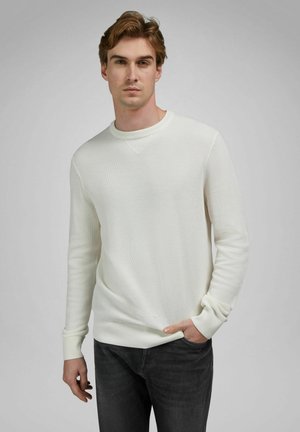 Joven de cabello castaño claro, vestido con un suéter blanco de textura y jeans gris oscuro, de pie con una mano en el bolsillo frente a un fondo gris.