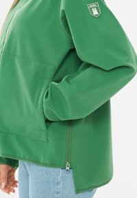 Veste verte avec main dans la poche, fermeture éclair latérale, et écusson du logo de la marque sur la manche gauche supérieure, portée avec un jean bleu clair.