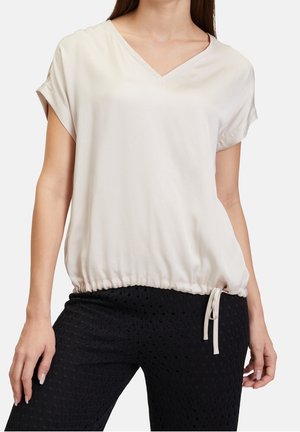 Donna che indossa una maglia beige larga con scollo a V e maniche corte, con orlo con coulisse abbinata a pantaloni neri strutturati.