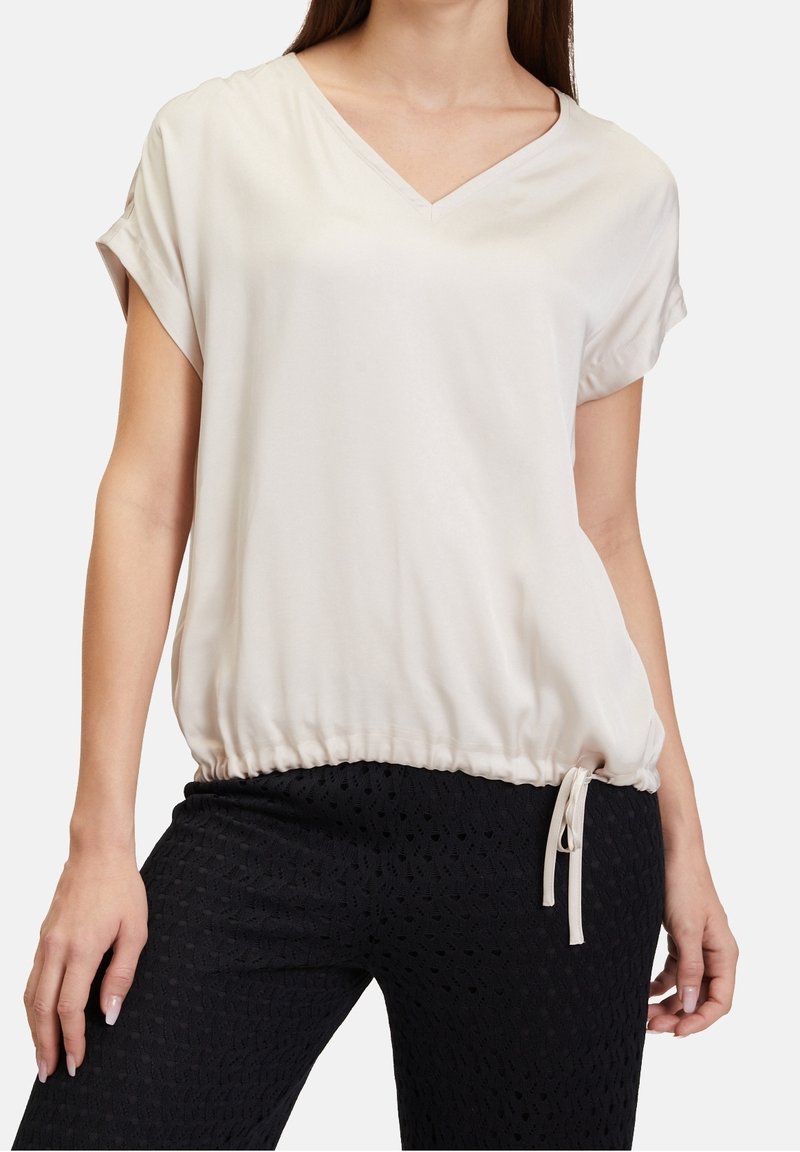 Femme portant un haut ample beige à manches courtes et col en V avec un ourlet à cordon, assorti à un pantalon noir texturé.
