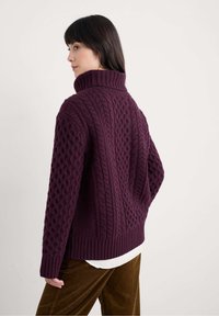 Übergroßer, tiefvioletter Pullover mit dickem Cable-Knit-Muster und hohem Rippkragen, kombiniert mit einem weißen Hemd und braunen Cordhosen.