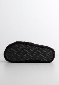 Semelle de pantoufle noire et duveteuse montrée à plat, texturée avec un motif géométrique répétitif incluant des flèches et des cercles pour l'adhérence.