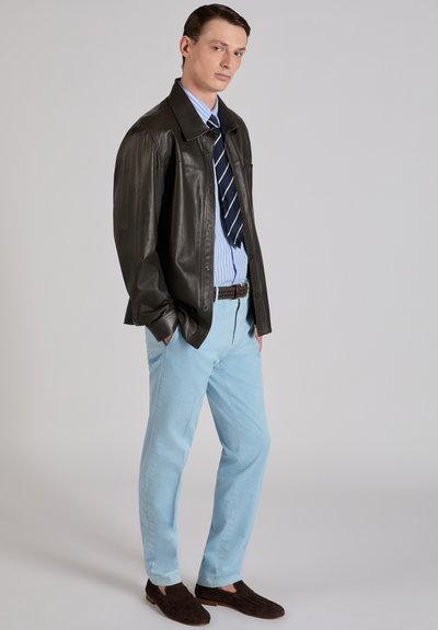 Mężczyzna w ciemnobrązowej skórzanej kurtce, niebiesko-białej koszuli w paski, granatowym krawacie w paski, jasnoniebieskich spodniach i ciemnobrązowych mokasynach, stojący z rękami w kieszeniach.