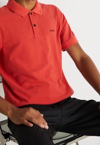 En person som sitter på en stol iförd en röd polo med "BOSS"-logga och svarta byxor, med händerna vilande på sina ben.