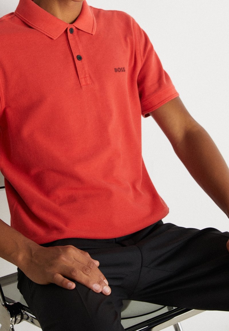 En person som sitter på en stol iförd en röd polo med "BOSS"-logga och svarta byxor, med händerna vilande på sina ben.