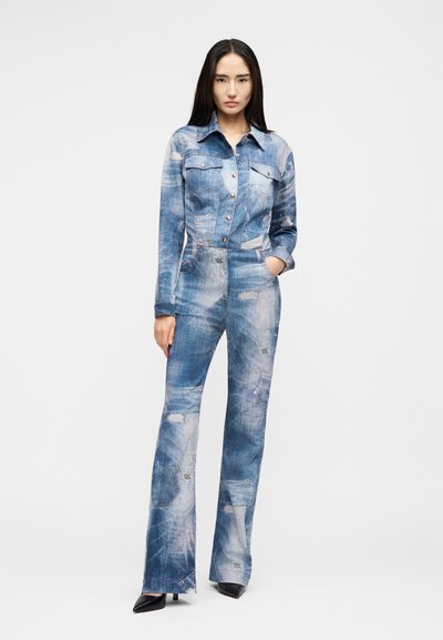 Γυναίκα με μακριά καστανά μαλλιά, φορώντας μπλε ολόσωμη φόρμα από τζιν με εφέ οξέος και μαύρες μυτερές γόβες, στέκεται με το ένα χέρι στην τσέπη.