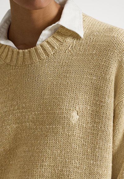 Bež pleten pulover z rebrastim izrezom in majhnim logotipom. Ima teksturirano, gosto tkanje in sproščen kroj.