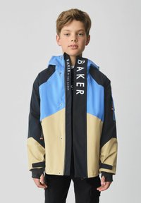 Farverig jakke med blå, sort og beige paneler, med hætte og sølvfarvede trykknapper. Inkluderer et fremtrædende mærkelabel på fronten.