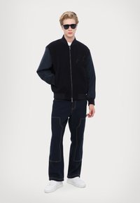 Giacca bomber navy con maniche a contrasto, chiusura lampo e logo testurizzato; abbinata a pantaloni blu scuro con cuciture visibili e sneaker bianche.